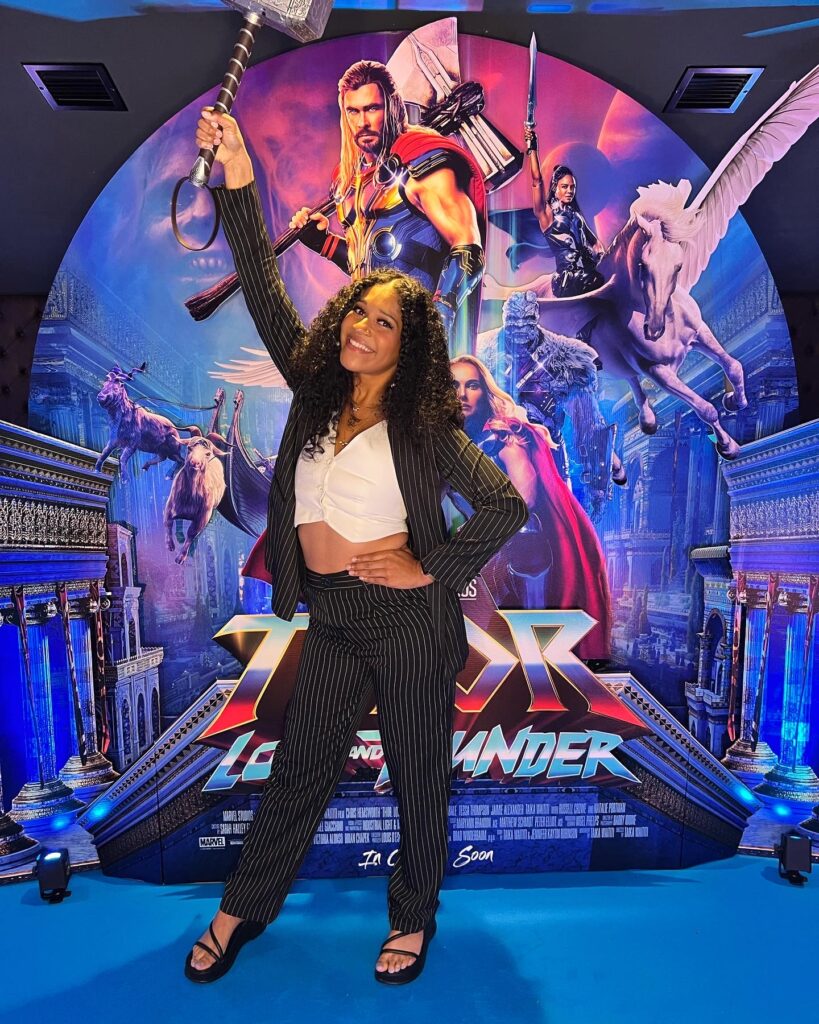 Dani at the media screening of Thor Love and Thunder in Leicester Square in front of a promotional poster form the film holding Thors hammer above her head and wearing a pink stripe suit.
